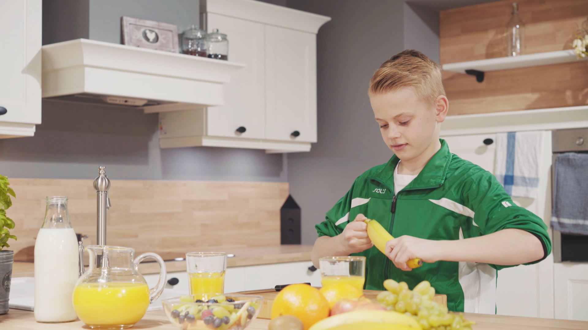 Ein Junge in grünem Trainingsanzug steht in einer Küche und presst eine Orange aus. Vor ihm auf der Arbeitsplatte stehen Gläser mit Orangensaft und eine Schüssel mit verschiedenen Früchten. Im Hintergrund sind Küchenschränke und eine Dunstabzugshaube zu sehen.
