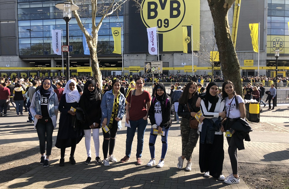 Eine Gruppe junger Frauen mit und ohne Kopftuch geht vor dem Signal Iduna Park, dem Stadion von Borussia Dortmund, vorbei. Im Hintergrund sind weitere Menschen und die Stadionfassade mit Werbebannern zu sehen.