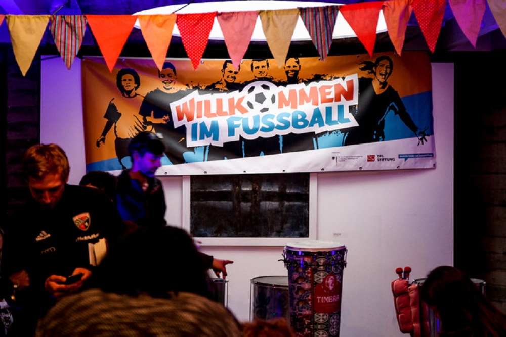 "Blick in ein Café mit buntem Banner 'Willkommen im Fußball' im Hintergrund, Teil des Bündnisses Ingolstadt der DFL Stiftung. Mehrere Personen sitzen an Tischen."