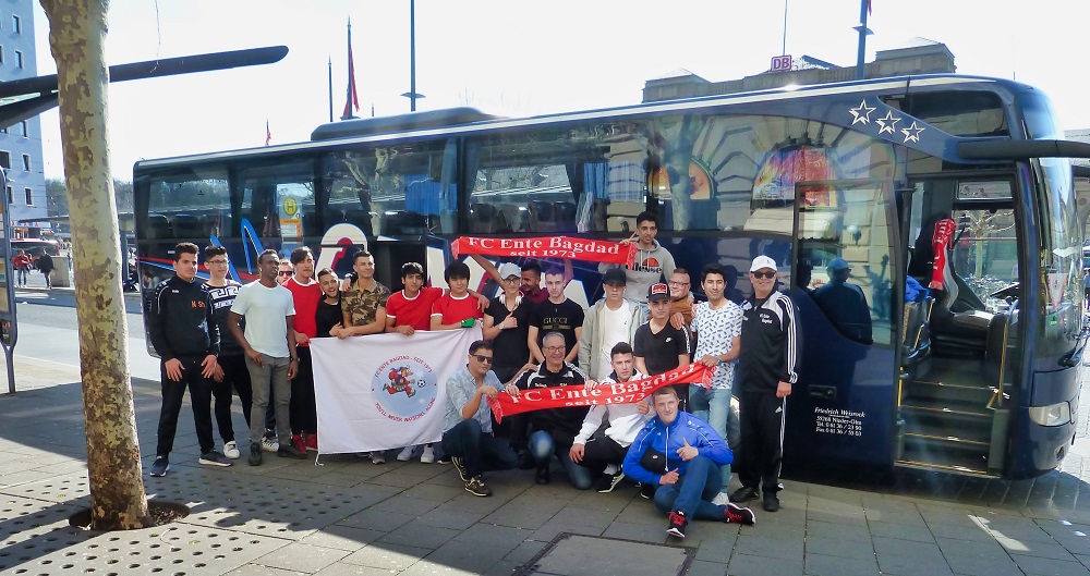 Eine Gruppe von Jugendlichen und jungen Erwachsenen steht vor einem Reisebus mit der Aufschrift "FC Ente Bagdad seit 1972". Im Hintergrund ist ein Bahnhof zu sehen.