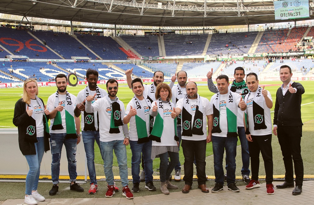 Eine Gruppe von Menschen steht auf dem Rasen eines Fußballstadions und hält weiße Banner mit dem Logo von Hannover 96 hoch. Im Hintergrund sind die leeren Zuschauerränge zu sehen.