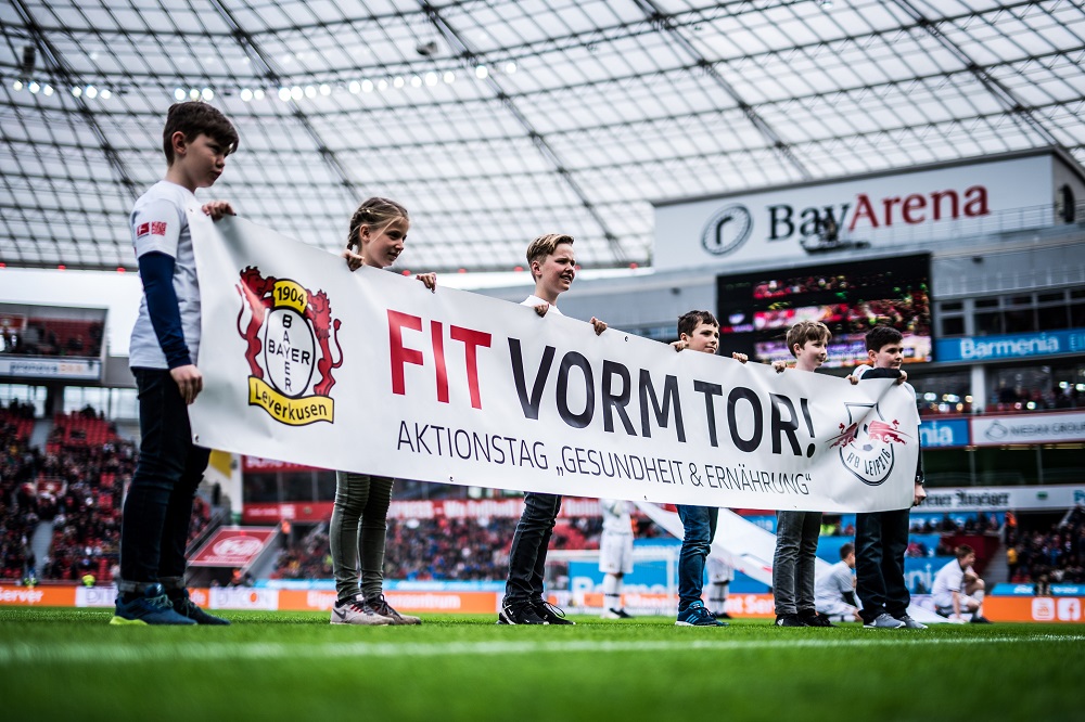 Sechs Kinder halten vor einem Fußballstadion ein großes Banner mit der Aufschrift "Fit Vorm Tor!" hoch. Im Hintergrund ist das Publikum und der Rasen des Stadions zu sehen.