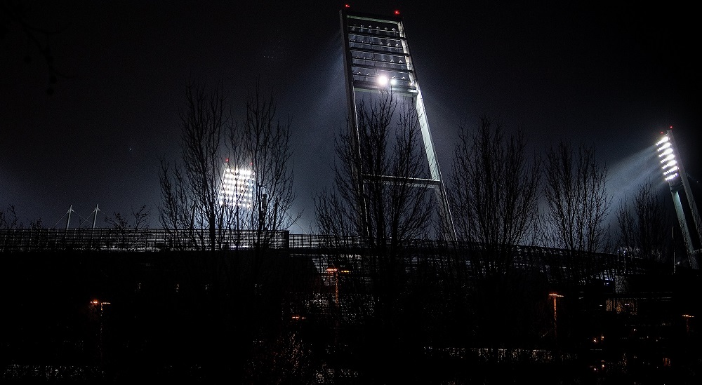 Nachtaufnahme eines Fußballstadions mit Flutlichtmasten, im Vordergrund eine dunkle Zuschauertribüne.