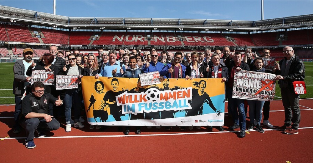 Eine große Gruppe von Menschen steht auf der roten Laufbahn eines Fußballstadions, im Hintergrund die Zuschauertribünen. Sie halten Banner mit der Aufschrift "Willkommen im Fußball" und "Vielfalt verbindet" hoch. Das Bild vermittelt ein Zeichen der Inklusion und Integration im Sport.