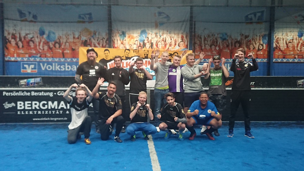 Eine Gruppe von Männern steht lächelnd in einer Sporthalle. Viele tragen T-Shirts mit Logos von Eintracht Braunschweig oder der DFL Stiftung. Im Hintergrund ist eine Werbetafel für "Bergmann Elektrizität" zu sehen.