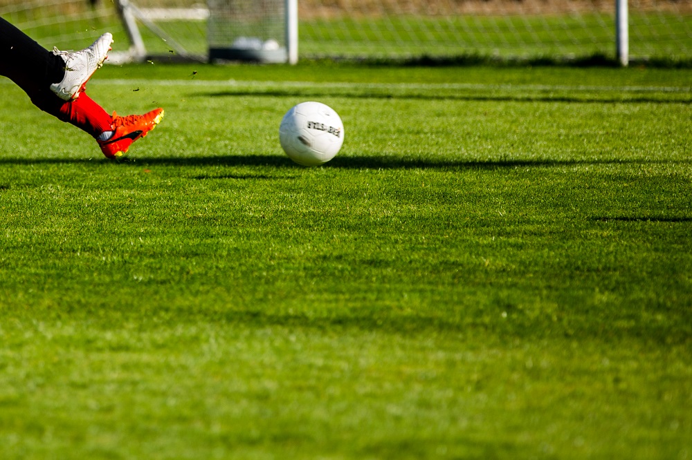 Ein Fußballspieler schießt den Ball auf ein Fußballtor, im Fokus liegt der Fuß des Spielers und der Ball. Im Hintergrund ist ein grünes Fußballfeld zu sehen.