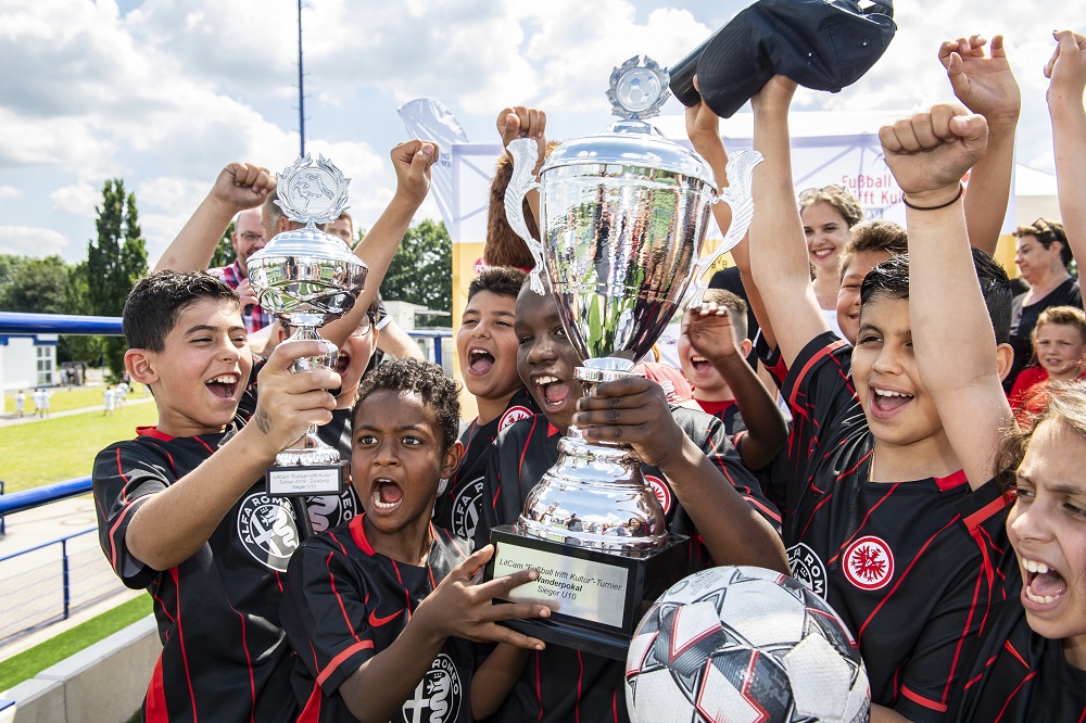 Jubelnde U10-Fußballmannschaft im roten Trikot, die gemeinsam einen großen Pokal in die Höhe stemmen. Im Hintergrund sind weitere Spieler und Zuschauer auf einem Fußballfeld zu sehen.
