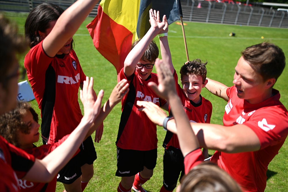 Jubelnde Kinder und Jugendliche in roten Trikots mit dem Logo von SC Freiburg feiern gemeinsam auf einem grünen Fußballplatz. Im Hintergrund ist eine Fahne zu sehen.