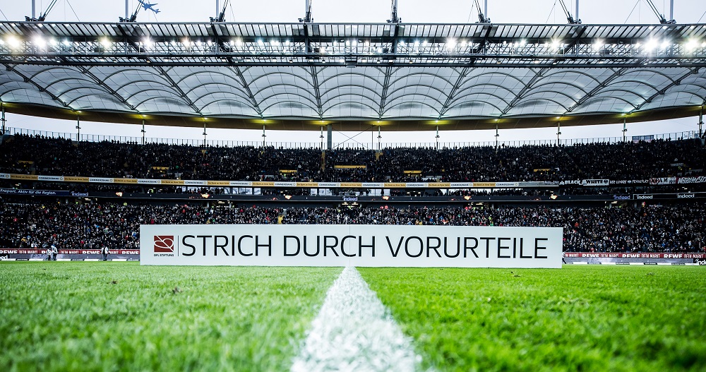 Fußballstadion gefüllt mit Zuschauern, auf dem Rasen ein Banner mit der Aufschrift "Strich durch Vorurteile".