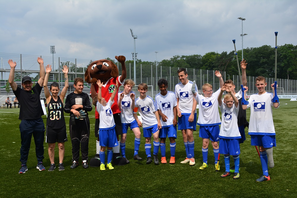 Eine Gruppe von Kindern in blauen Fußballtrikots und roten Shorts steht auf einem grünen Fußballfeld. Neben ihnen steht ein Mann in schwarzer Sportkleidung und eine Fuchs-Maskottchenfigur. Alle heben die Hände in die Luft und lächeln in die Kamera. Im Hintergrund sind grüne Büsche und ein Fußballtor zu sehen.