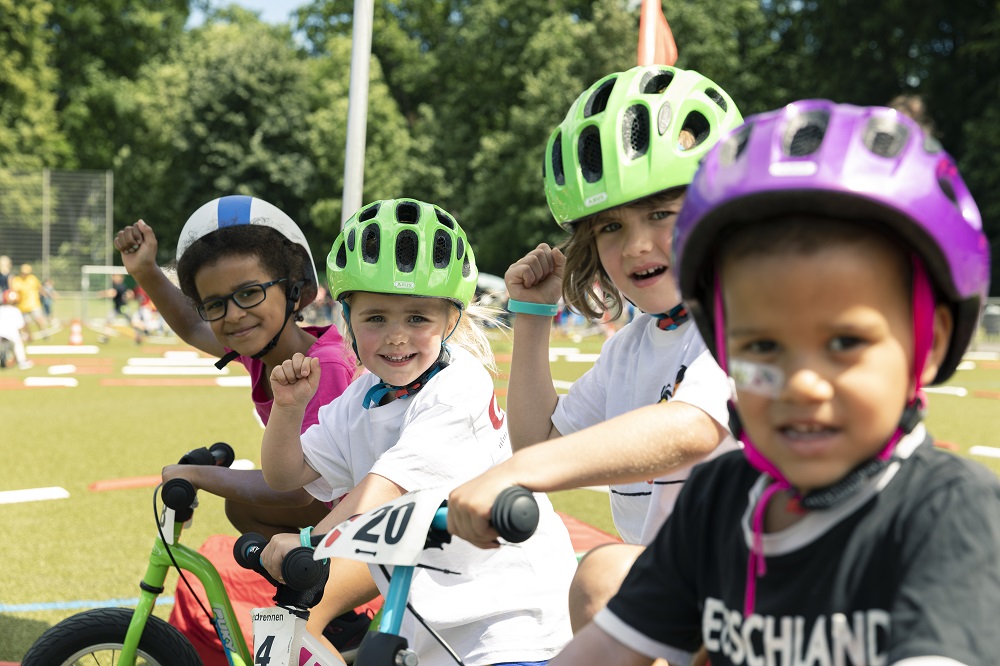 Eine Gruppe von Kindern mit Fahrrädern und Helmen feiert auf einem Fußballplatz. Die Kinder schauen mit hoch erhobenen Armen und strahlenden Gesichtern in die Kamera. Im Hintergrund ist grünes Gras und ein Fußballtor zu sehen.
