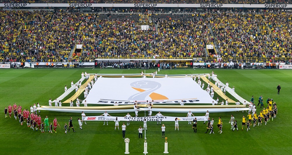 Auf dem Spielfeld des Dortmunder Stadions ist neben dem Supercup-Logo ein Banner mit der Aufschrift "Spitzensport verbindet" und den Logos von DFL Stiftung und Deutscher Sporthilfe zu sehen. Vor diesem Banner stehen drei Personen hinter den drei Trophäen des deutschen Fußballs, der Meisterschale, dem Supercup und dem DFB-Pokal.