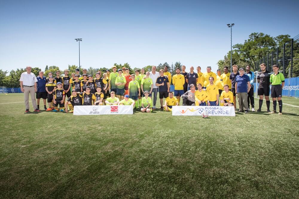 Eine große Gruppe von Menschen steht auf einem Fußballfeld zusammen und lächelt in die Kamera. Viele tragen gelbe T-Shirts mit dem Aufdruck "FußballFreunde". Im Hintergrund sind Bäume und eine Sportanlage zu sehen.