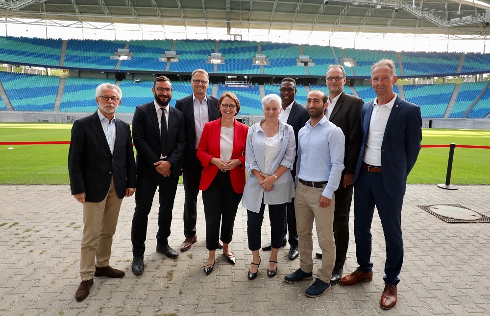 Eine Gruppe von sieben Personen steht vor dem Fußballstadion. Im Zentrum steht Annette Widmann-Mauz, erkennbar an ihrem roten Blazer, und schaut in die Kamera. Die anderen Personen stehen seitlich von ihr und lächeln ebenfalls in die Kamera. Im Hintergrund ist das Spielfeld und die Zuschauertribüne des Stadions zu sehen.