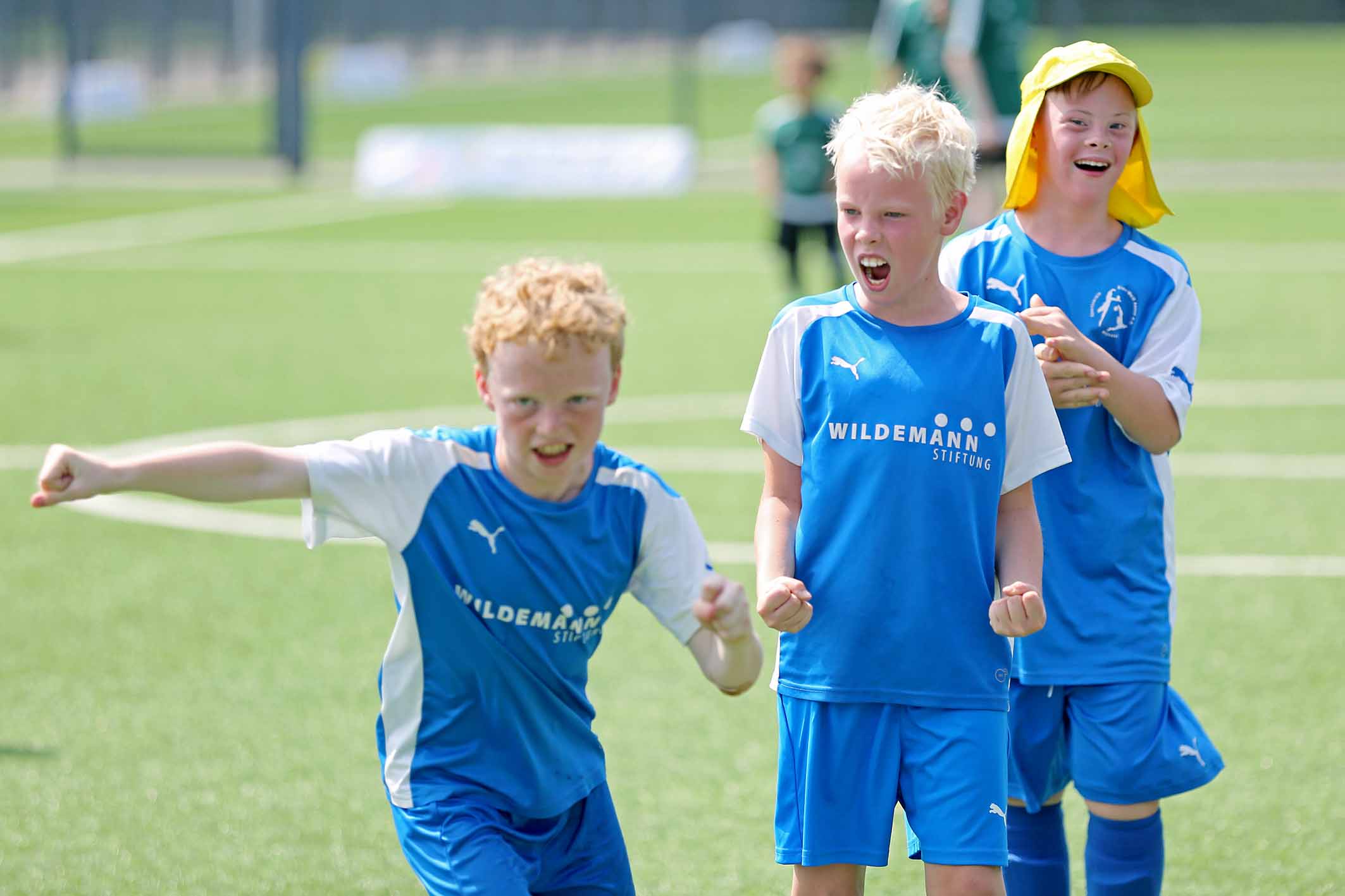 Jubelnde Kinder in blauen Trikots mit dem Aufdruck "WILDEMANN STIFTUNG" auf einem Fußballfeld.