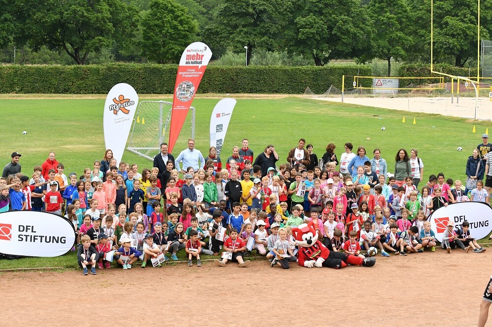 Eine große Gruppe von Kindern und Jugendlichen steht auf einem Fußballplatz vor einem Tor und lächelt in die Kamera. Im Hintergrund sind Fahnen mit dem Logo der DFL Stiftung und des "fit4future"-Cups zu sehen.