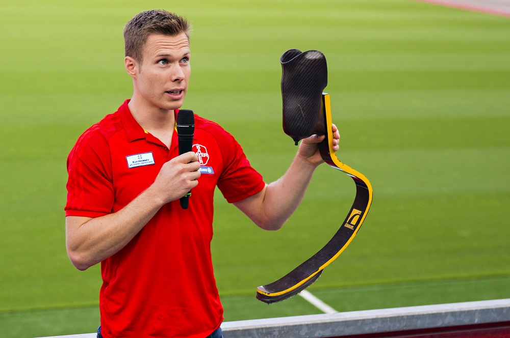Markus Rehm, Paralympics-Sprinter, hält seine Prothese in die Höhe und spricht in ein Handmikrofon. Im Hintergrund ist ein Fußballfeld zu sehen.