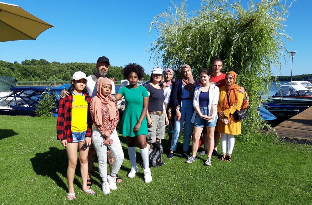 Eine Gruppe von jungen Menschen steht lächelnd auf einer grünen Wiese am Ufer eines Sees. Im Hintergrund sind Bäume und Boote zu sehen. Es handelt sich um eine Aufnahme vom "Fußball Bündnis Berlin-Köpenick" der DFL Stiftung.