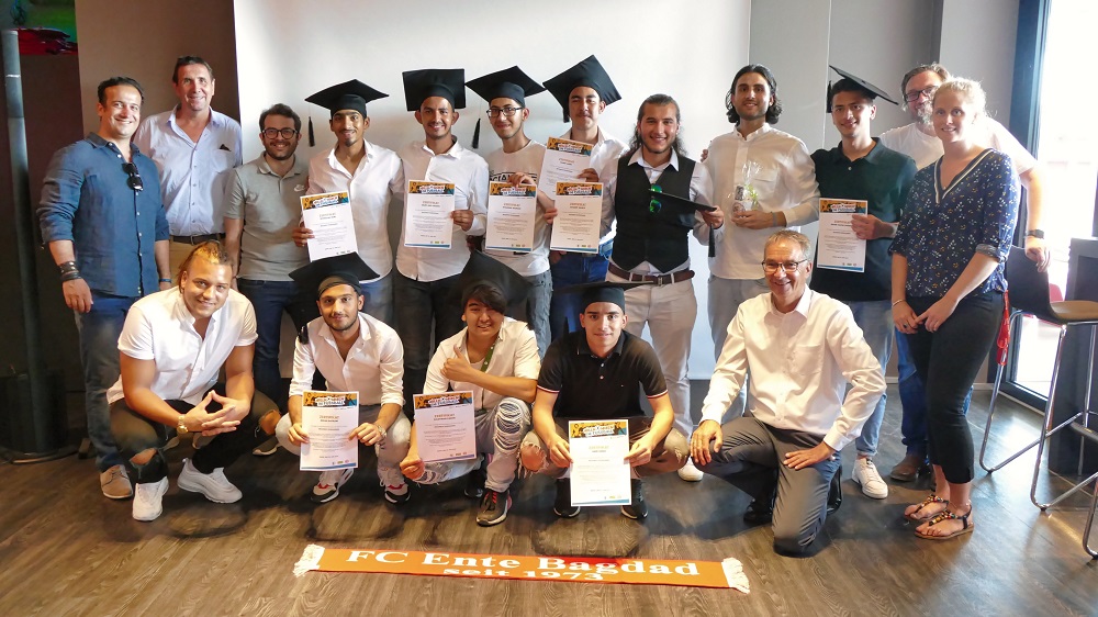 Eine Gruppe junger Männer, vermutlich Fußballspieler des FC Ente Bagdad, posiert mit ihren Zertifikaten. Einige tragen dabei noch ihre Abschlusskappen. Im Hintergrund stehen weitere Personen, möglicherweise Trainer oder Betreuer. Vor der Gruppe liegt ein Banner mit der Aufschrift "FC Ente Bagdad seit 1973".