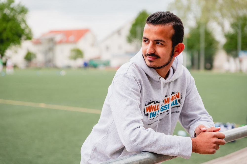 Junge Person mit dunklen Haaren und grauem Hoodie, auf dem "Willkommen im Fußball" steht, blickt lächelnd in die Ferne, im Hintergrund ein Fußballfeld.