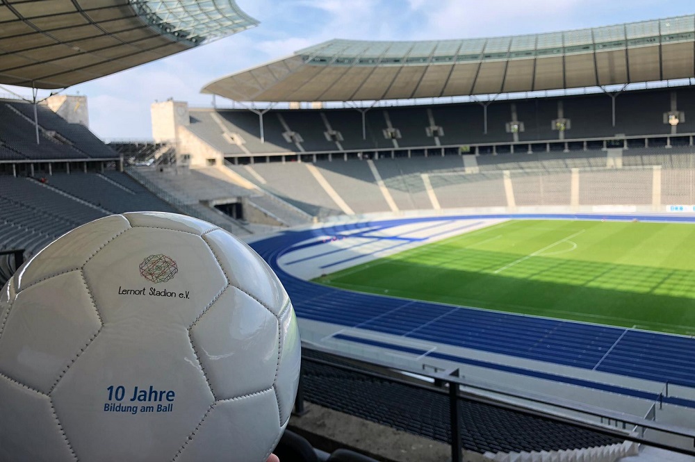 Ein Fußball liegt im Vordergrund, vor dem Hintergrund eines Fußballstadions mit leeren Zuschauerrängen. Auf dem Ball ist das Logo "Lernort Stadion e.V." zu sehen. Im linken unteren Bildrand ist ein Logo mit der Aufschrift "10 Jahre Bildung am Ball" zu sehen.