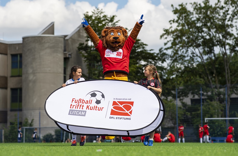 Ein Maskottchen der DFL Stiftung steht auf einem Banner mit der Aufschrift "Fußball trifft Kultur" vor einer Gruppe von jungen Fußballspielerinnen. Im Hintergrund ist ein Gebäude zu sehen.