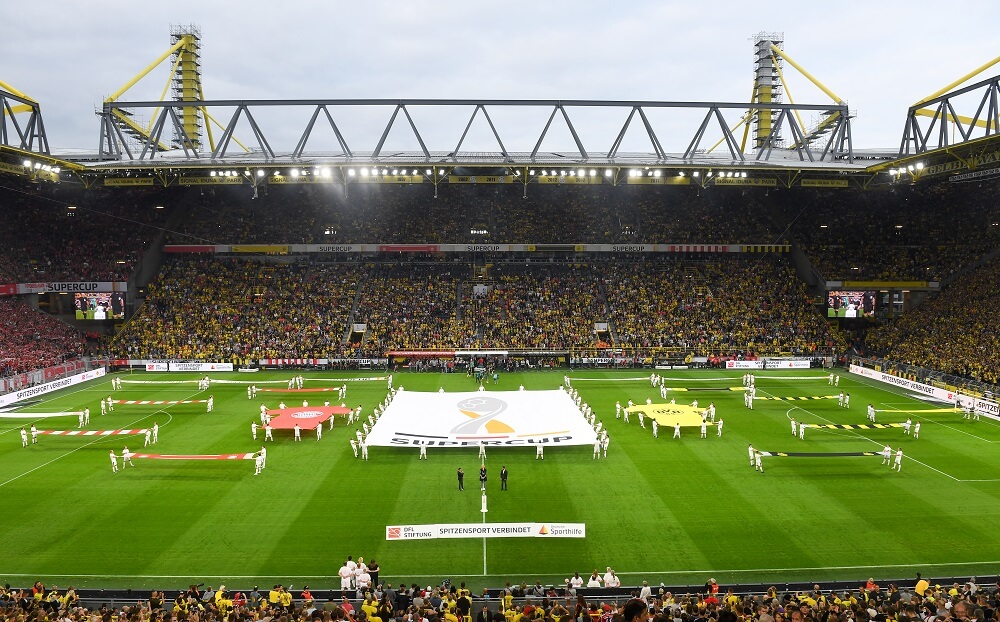 Fußballstadion gefüllt mit Zuschauern, auf dem Rasen sind Menschen mit einer großen Banner zu sehen, auf dem "SUPER CUP" steht.