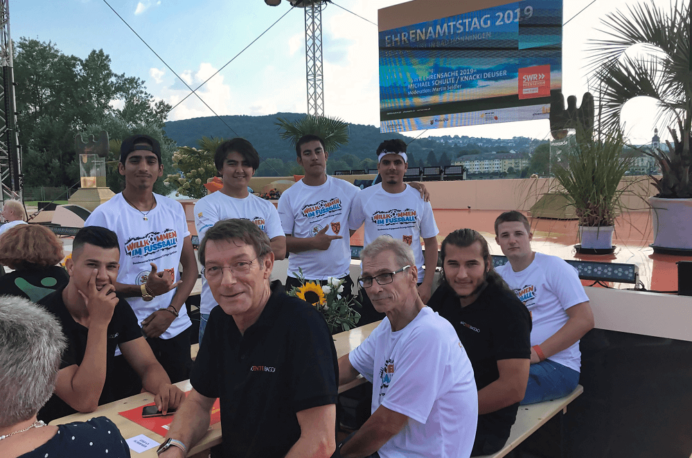 Eine Gruppe Jugendlicher im Trikot des "FC Ente Bagdad" posiert mit einem Mann im blauen Polo vor einer Leinwand mit dem Aufdruck "EHRENSACHE". Im Hintergrund eine Hügellandschaft und ein grauer Himmel.