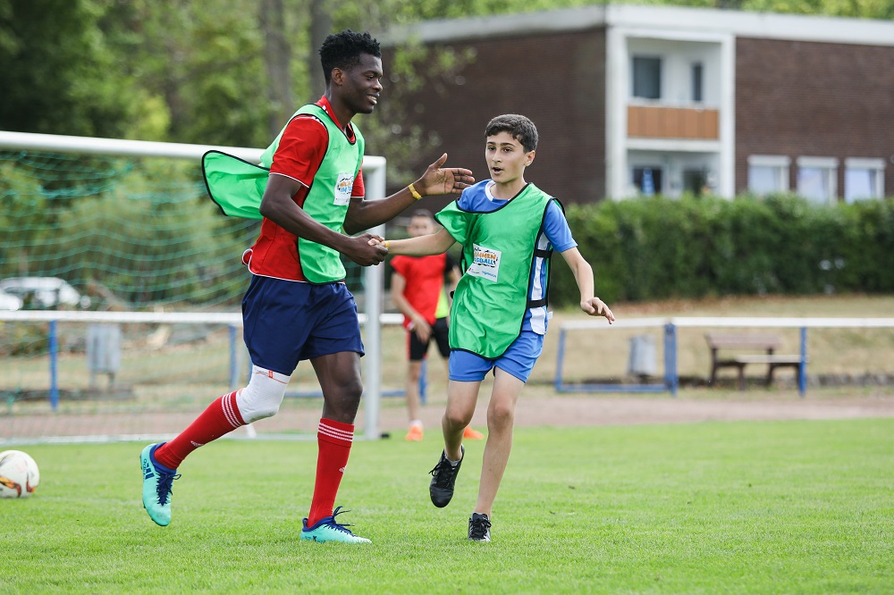 Zwei jugendliche Fußballspieler rennen auf einem grünen Rasenplatz. Der Spieler links trägt ein rotes und schwarzes Trikot mit Trainingsweste, der Spieler rechts ein grünes Trikot mit Trainingsweste. Im Hintergrund ist ein Fußballtor und Gebäude zu sehen.