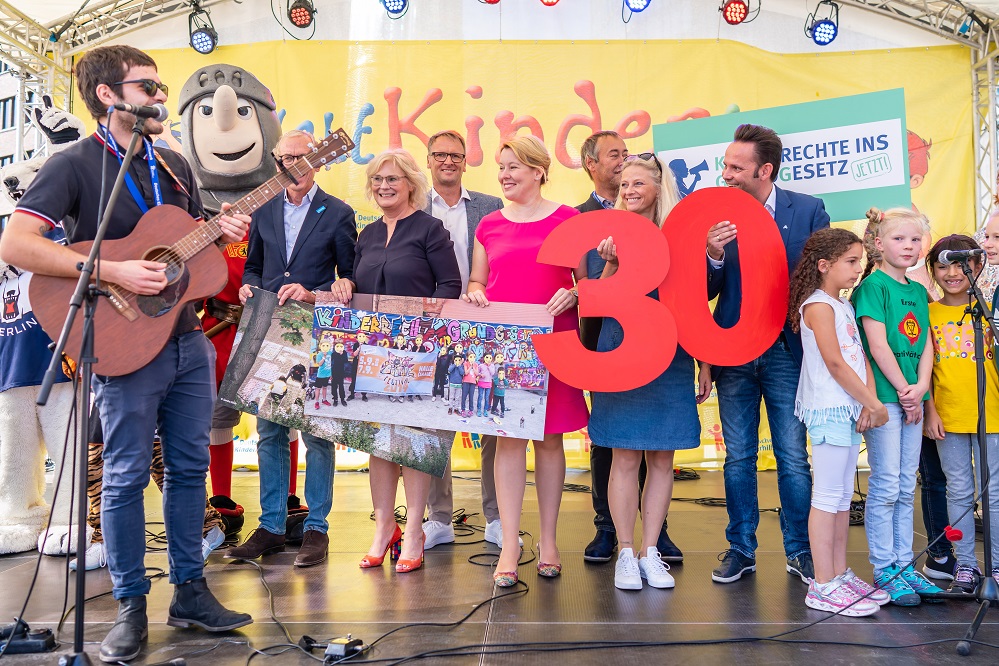 Eine Gruppe von Menschen, darunter Politiker und Kinder, steht auf einer Bühne vor einem Banner mit der Aufschrift "30". Ein Mann spielt Gitarre, während ein anderes Kind ein Plakat mit bunten Zeichnungen hält. Im Hintergrund ist ein Banner mit der Aufschrift "Rechte Gesetz" zu sehen.