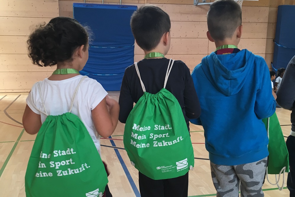 Three children (one girl and two boys) are standing in a gym with their backs to the camera. Two of the children are carrying gym bags with the inscription 