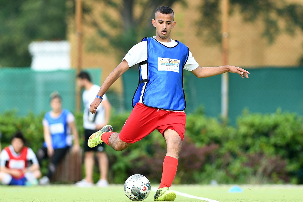 Ein junger Fußballspieler mit roter Shorts und blauem Trainingsshirt dribbelt auf einem grünen Rasen. Im Hintergrund sind weitere Spieler zu sehen.