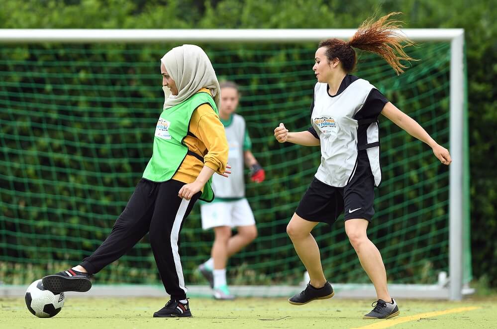 Zwei junge Frauen mit Kopftuch und ohne, in Sportkleidung und Westen, spielen Fußball auf einem grünen Kunstrasenplatz. Im Hintergrund ist ein Fußballtor und eine weitere Spielerin erkennbar.