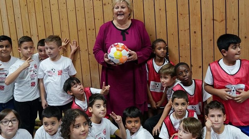 Eine Frau hält einen Fußball vor einer Gruppe lachender Kinder in Sportkleidung. Im Hintergrund eine Holzvertäfelung.