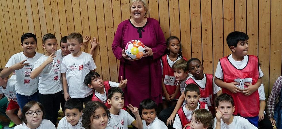 Eine Frau hält einen Fußball vor einer Gruppe lachender Kinder in Sportkleidung. Im Hintergrund eine Holzvertäfelung.