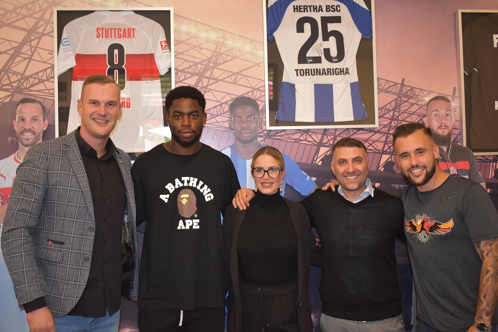 Eine Gruppe von sechs Personen steht vor einer Wand mit Fußballmotiven. Im Hintergrund sind Poster von Spielern und das Logo von Hertha BSC zu sehen. Die Gruppe besteht aus drei Männern und zwei Frauen. Ein Mann trägt ein rotes T-Shirt, ein anderer ein schwarzes T-Shirt mit einem Logo. Eine Frau trägt eine Brille und ein schwarzes Oberteil. Die anderen Personen sind ebenfalls lässig gekleidet.