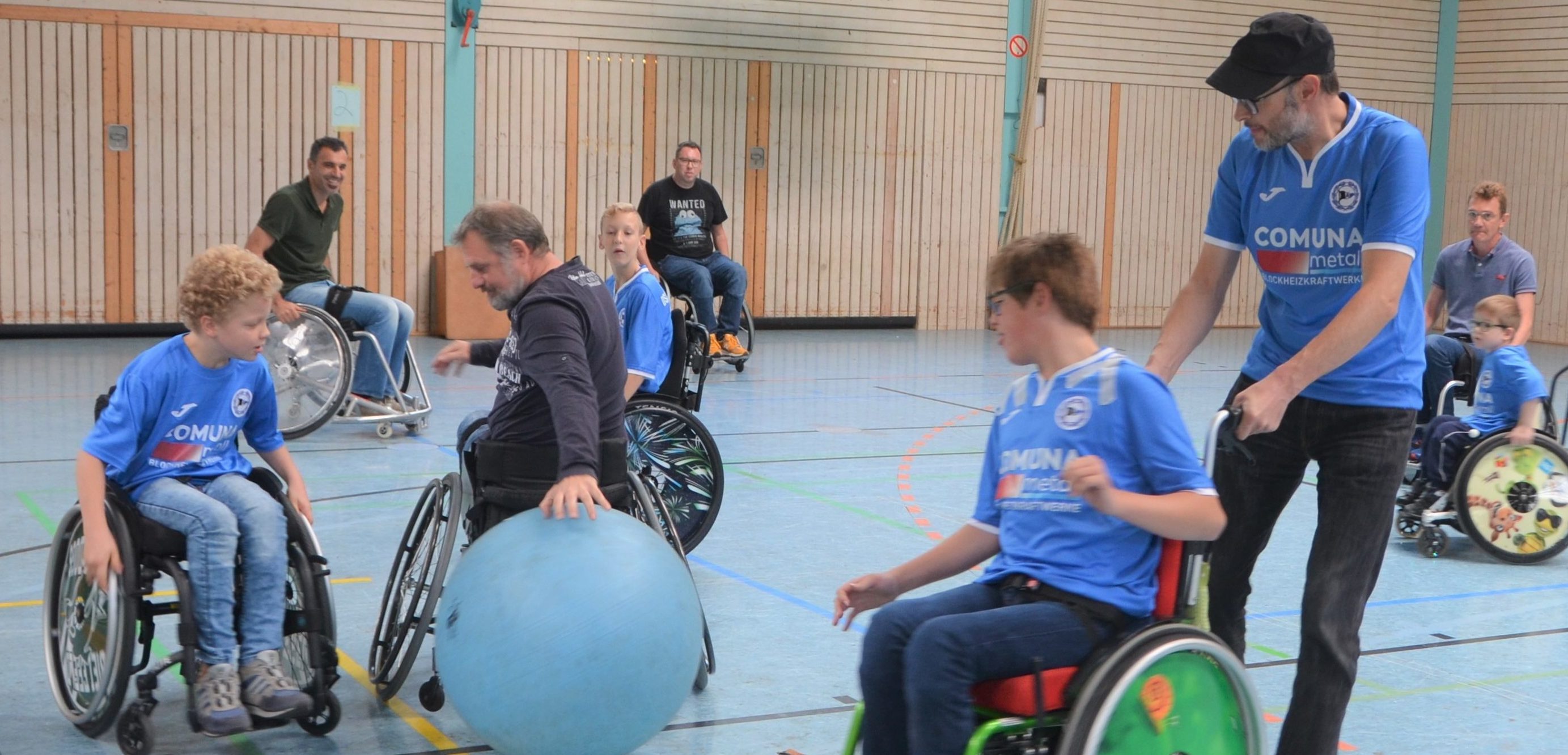 Rollstuhl-Fußballturnier der RBG Dortmund 51: Mehrere Spieler im Rollstuhl auf dem Spielfeld, einige konzentrieren sich auf den blauen Ball, während andere pausieren oder sich austauschen. Im Hintergrund sind weitere Personen und die Hallenwand zu sehen.
