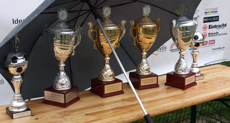 Pokale für die Gewinner der Bolzplatzliga U15 und U18 stehen auf einer grünen Fläche, teilweise im Schatten eines Regenschirms. Im Hintergrund ist ein Banner mit dem Logo des Sportkreises Frankfurt zu sehen.