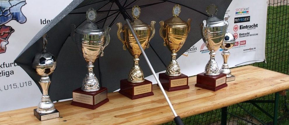 Pokale für die Gewinner der Bolzplatzliga U15 und U18 stehen auf einer grünen Fläche, teilweise im Schatten eines Regenschirms. Im Hintergrund ist ein Banner mit dem Logo des Sportkreises Frankfurt zu sehen.
