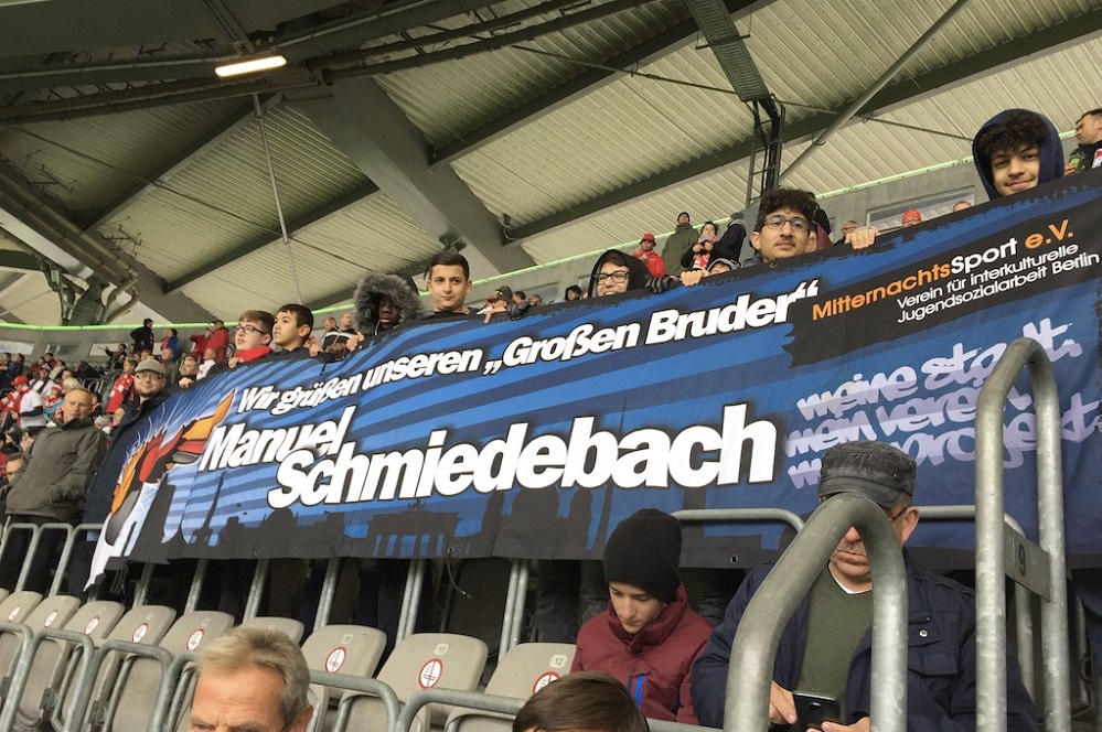 Banner mit der Aufschrift "Wir grüßen unseren ‚Großen Bruder‘ Manuel Schmiedebach" im Stadion während eines Fußballspiels. Im Hintergrund sind Zuschauerreihen zu sehen.