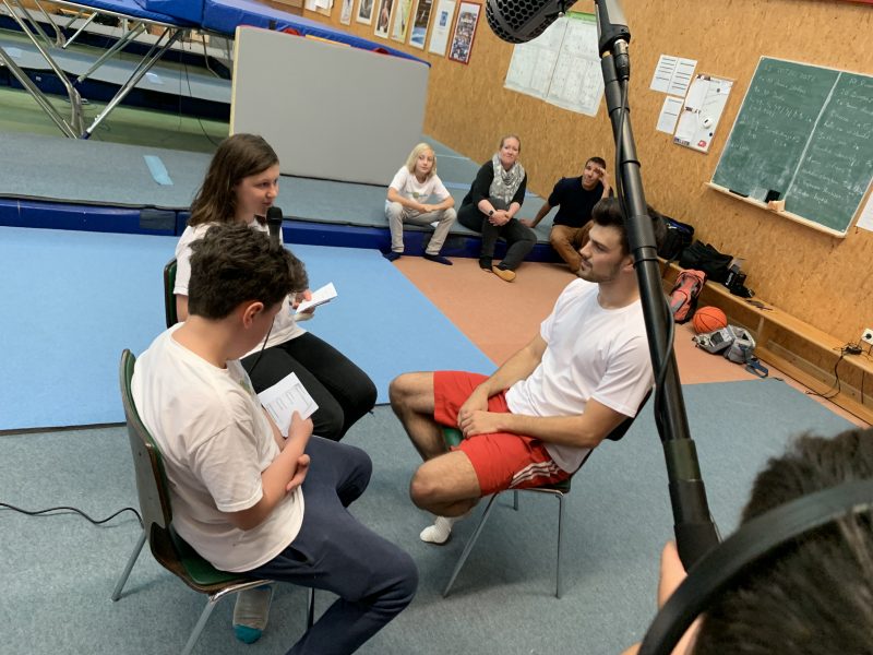 Eine Gruppe junger Menschen sitzt in einem Turnraum in einem Kreis auf Stühlen und nimmt an einem Quiz teil. Ein Mann hält eine Karte mit einer Frage hoch, während die anderen Teilnehmer aufmerksam zuhören und überlegen. Im Hintergrund sind eine Tafel und Sportgeräte zu sehen.