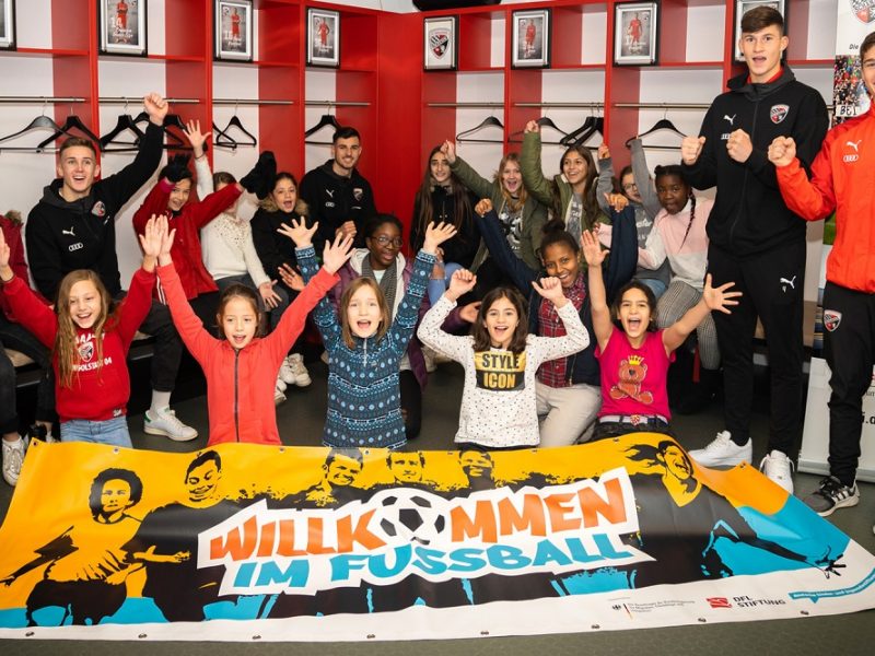 Eine Gruppe von Kindern und Jugendlichen steht mit hoch erhobenen Armen und jubelnd vor einer großen, bemalten Leinwand mit der Aufschrift "Willkommen im Fußball". Im Hintergrund stehen mehrere Erwachsene, darunter vermutlich Fußballspieler. Die Szene wirkt fröhlich und einladend, die Leinwand deutet auf eine Aktion der DFL Stiftung hin.