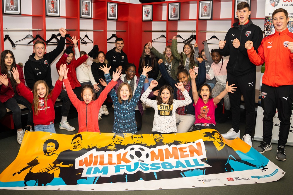 Eine Gruppe von Kindern und Jugendlichen steht mit hoch erhobenen Armen und jubelnd vor einer großen, bemalten Leinwand mit der Aufschrift "Willkommen im Fußball". Im Hintergrund stehen mehrere Erwachsene, darunter vermutlich Fußballspieler. Die Szene wirkt fröhlich und einladend, die Leinwand deutet auf eine Aktion der DFL Stiftung hin.