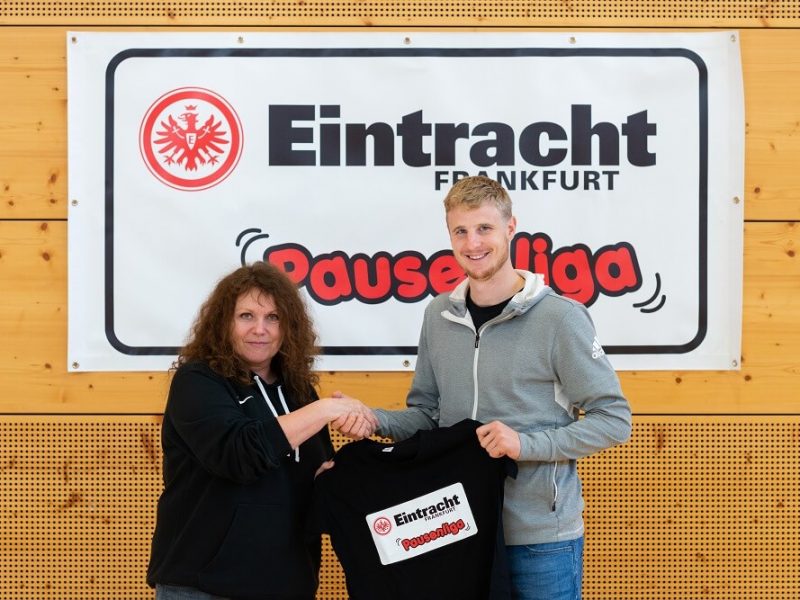 Ein Mann mit blonden Haaren und grauem Hoodie schüttet die Hand einer Frau. Im Hintergrund ein Banner mit dem Logo von Eintracht Frankfurt und dem Schriftzug "Pausenliga".