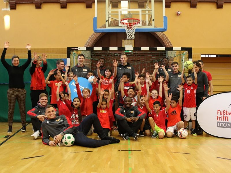 Gruppe von Kindern und Jugendlichen in roten Trikots, umgeben von Betreuern und DFL Stiftung Mitarbeitern, jubelt in einer Sporthalle. Im Hintergrund ein Basketballkorb und ein Banner mit dem Aufdruck "Fußball trifft Kultur LITCAM".
