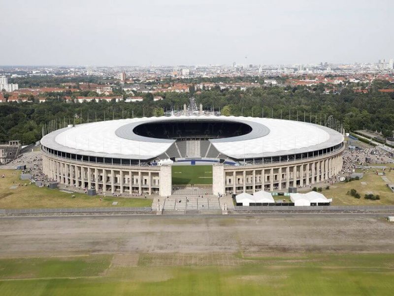 Das Olympiastadion Berlin aus der Vogelperspektive, umgeben von Grünflächen und mit der Skyline der Stadt im Hintergrund.