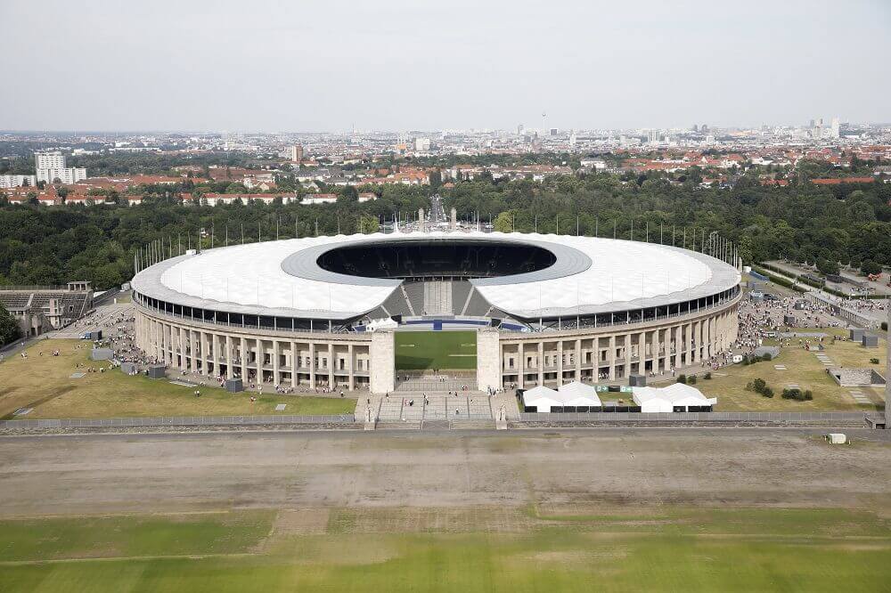 Das Olympiastadion Berlin aus der Vogelperspektive, umgeben von Grünflächen und mit der Skyline der Stadt im Hintergrund.