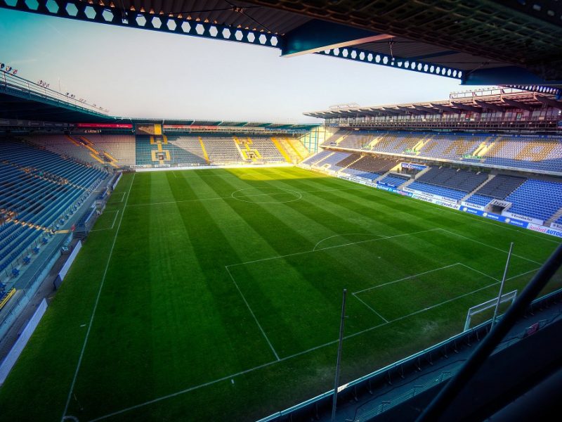 Fußballstadion mit grüner Rasenfläche und blauen Sitzen, Blick von oben aus einer Tribüne.