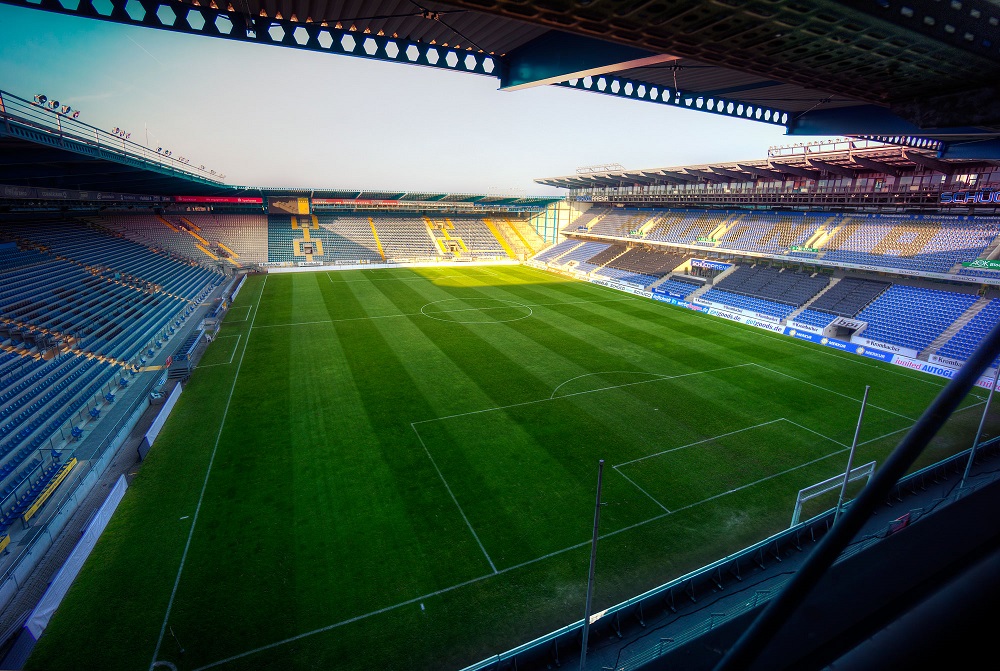 Fußballstadion mit grüner Rasenfläche und blauen Sitzen, Blick von oben aus einer Tribüne.
