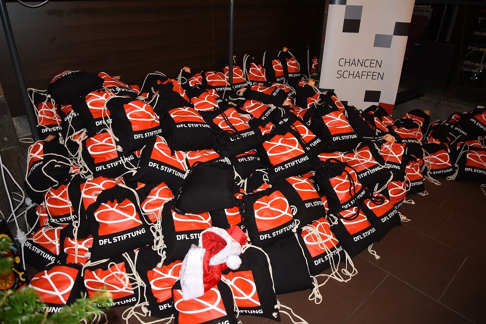 Stapelweise rote Schals mit dem Aufdruck "DFL STIFTUNG" liegen auf einem Tisch, vermutlich als Gastgeschenke bei einem Weihnachtsessen. Im Hintergrund ist ein Schild mit der Aufschrift "CHANCEN SCHAFFEN" zu sehen.